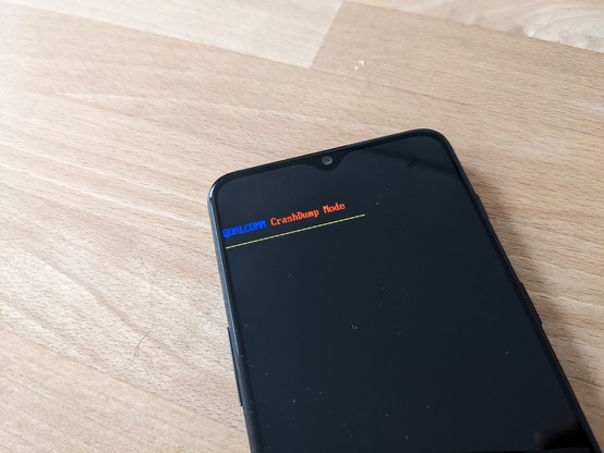 Photo of a smartphone on a wooden table surface. The screen is showing: "QUALCOMM CrashDump Mode" in blue, red and yellow letters.