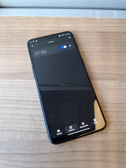 Photo of a OnePlus 6T on a wooden surface with the Phosh UI. The Clocks app is openend, listing an alarm at 7 o'clock.