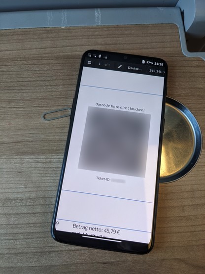 Photo of a OnePlus 6T on a wooden surface with the Phosh UI. The document viewer app is openend, showing a train ticket with a QR code in the middle. The QR code and the ticket ID have been blurred. Next to the phone is a SIM eject pin.