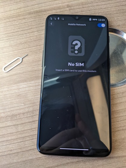 Photo of a OnePlus 6T on a wooden surface with the Phosh UI. The mobile network settings are open, showing the text "No SIM; Insert a SIM card to use this modem". Next to the phone is a SIM eject pin.