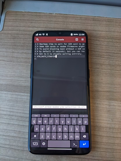 Photo of a OnePlus 6T on a wooden surface with the Phosh UI. The terminal app is openend, showing a text editor. The text editor is displaying a configuration file. The current, highlighted line reads "sim_wait_time=30"