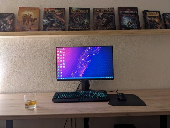 A photo of a desk with a computer monitor in the center. On the desk are also a mouse and keyboard, a mousepad and a glass of whiskey. The computer is running KDE Plasma and about a dozen of game icons can be seen. Above the desk is a bookshelf presenting a handful of tabletop RPG rulebooks.